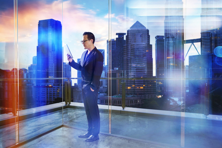 Businessman use digital device stand at modern transparent office with beautiful city skyline , evening scene .の写真素材