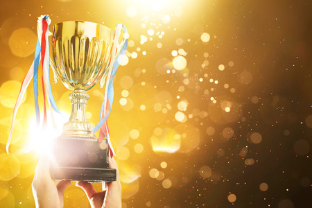 Hand raised, holding gold cup with abstract shiny lights . award and victory concept .の写真素材