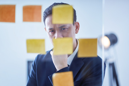 Asian pensive businessman  looking at glass wall with sticker notes , brainstorming concept .の写真素材