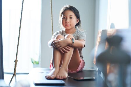 Pretty asian little girl black hair in grey shirt sitting on living room sofa and feeling happyの写真素材