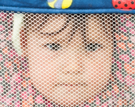 One year old half Chinese half caucasian baby hanging in a play pen looking through mesh.の写真素材