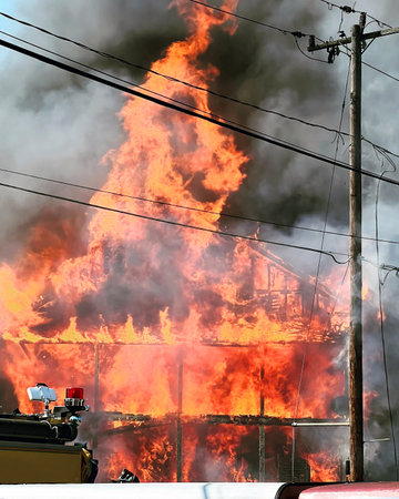 Photo of a raging, full flame house fire with thick gray and black smoke rising from it. Part of a fire truck can be seen in the foreground with a red light. Black smoke lingers.の写真素材