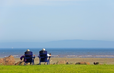 Two people sit on blue Adirondack beach chairs overlooking the beach.の写真素材