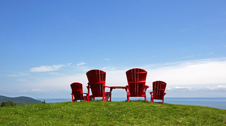 Four red Adirondack chairs on a grassy slope overlooking a body of water.の写真素材