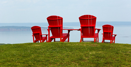 Four red Adirondack chairs on a grassy slope overlooking a body of water.の写真素材