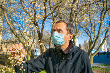 The man in the protective mask in a park near the flowering magnolia tree wants to smell flowers on...