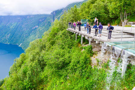 Geiranger, Norway - Jyne 26, 2019: Tourists on Eagles Road viewpoint, near water falling from the platform. Landscape of Geirangerfjord and Seven Sisters Waterfall in the background. Summer travel.のeditorial素材