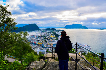 Tourist man looks on the Alesund cityscape from Aksla viewpoint. Neoclassical and neo-Gothic stone buildings. Art Nouveau architecture. Sun rays illuminate Atlantic Ocean and islands. Norway.の写真素材