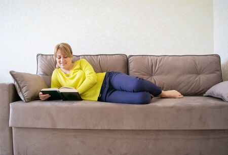 Caucasian woman is reading a book lying at home on the comfortable sofa ones during quarantine and social distance at Coronovirus (COVID-19) pandemic. Home leisure. It's good to be home.の写真素材