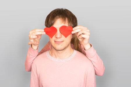 Girl stands behind young, caucasian guy and holds two red hearts in front of him, symbols of love. Couple of lovers, young, smiling, beautiful, light skinned caucasian people on light gray background.の写真素材