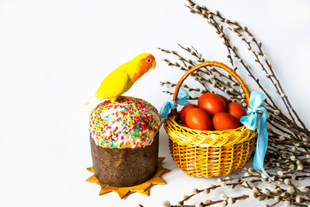 Easter cake, a wicker basket with brown Easter eggs, willow twigs and a lovebird parrot of bright festive yellow and red colors on a white background. Easter greeting card. Copy space.の写真素材