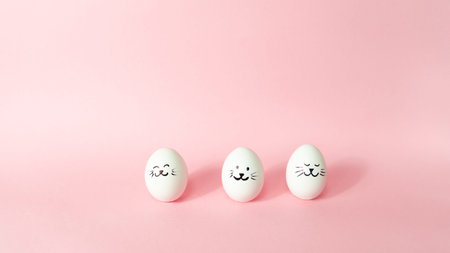 Three white chicken eggs with painted cute bunny faces are isolated on a light pink pastel...