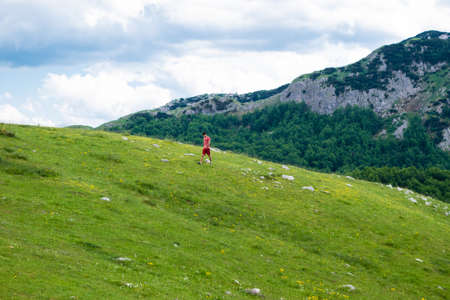 Guy tourist walks on green hill. Durmitor National Park. Highest road pass in Montenegro, located in north of Montenegro. Summer cloudy landscape.の写真素材