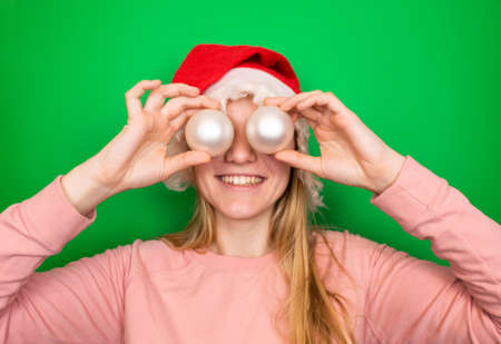 Young smiling blonde caucasian girl with long hair in red winter Christmas Santa Claus cap holds Christmas decorative white balls in her hands near her eyes. Green background with copy space.の写真素材