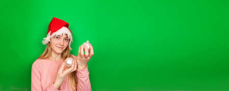 Young smiling blonde light skinned caucasian girl with long hair in a red winter Christmas Santa Claus cap holds Christmas tree decorative white balls in her hands. Green background with copy space.の写真素材