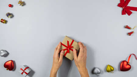 Woman hands hold a box with a gift tied with a red ribbon in a festive frame on a light gray background. Red hearts, bow and other decorations with copy space.の写真素材