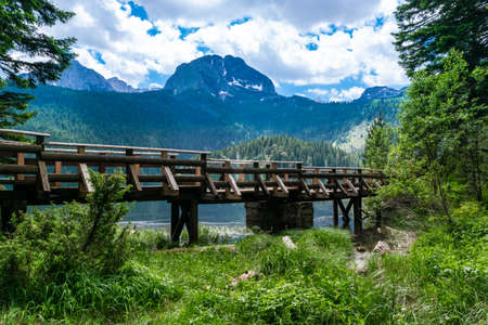 Wooden footbridge over stream against background of Meded Peak and Glacial Black Lake. Durmitor National Park. Walking path near lake is a popular destination for recreation and hiking. Montenegro.の写真素材
