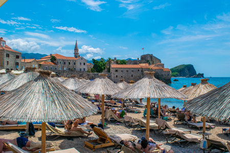 Budva, Montenegro - July 4, 2021: Resting people on sun loungers under thatched umbrellas on Brijeg od Budva beach on background of old city walls fortress.のeditorial素材