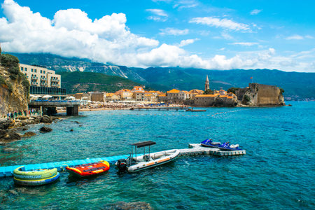 Budva, Montenegro - July 4, 2021: Pontoon dock with scooters on background of beach and old city walls fortress. Budva Old Town. Beautiful blue summer sunny seascape.のeditorial素材