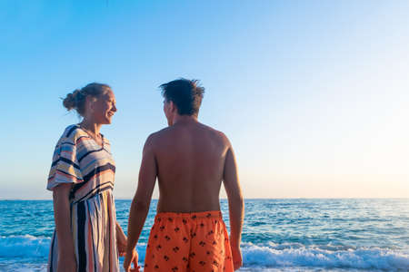 Young joyful girl with blond hair with guy in the rays of sunset on the beach against the backdrop of the sea and a cloudless blue sky. Summer rest.の写真素材