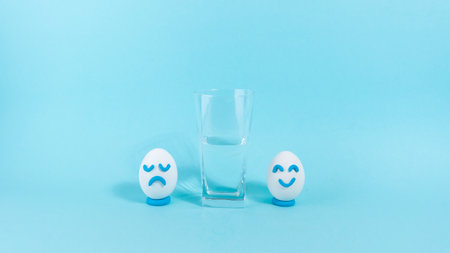 Next to the half-filled glass, two eggs with a cheerful and sad expression. Concept of optimist and pessimist. blue background. Copy space.の写真素材