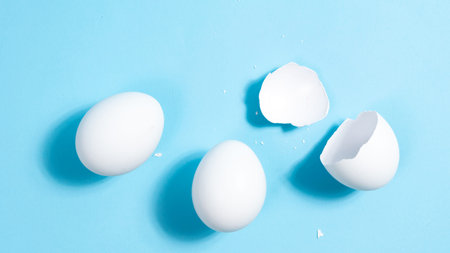 Two whole white chicken eggs and one broken on a blue background. The concept of protein nutrition...