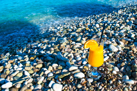 Fresh orange juice in a glass with a straw and an orange slice on a pebble beach against the backdrop of a clear calm blue sea. Summer holiday concept.の写真素材