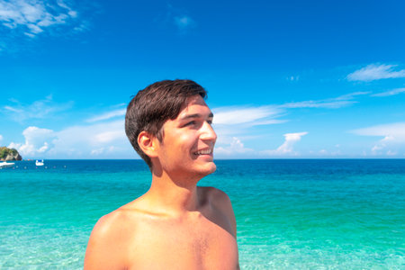 Young tanned smiling guy on the beach against a blue beautiful clean sea and sky. Concept of summer vacation by the sea, getting vitamin D while taking morning sunbaths.の写真素材