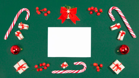 Christmas greeting card. Rectangular frame of candy canes, christmas balls, twigs with red berries and gifts boxes on viridian green background. White sheet of paper in center. Template for text.の写真素材