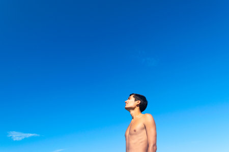 Close-up figure of a young tanned guy on the beach against the blue sky. The concept of a summer vacation by the sea, getting vitamin D while taking morning sunbathsの写真素材