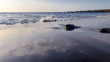 Surf waves close-up on a beach with black volcanic sand illuminated by the sun. Stone from...