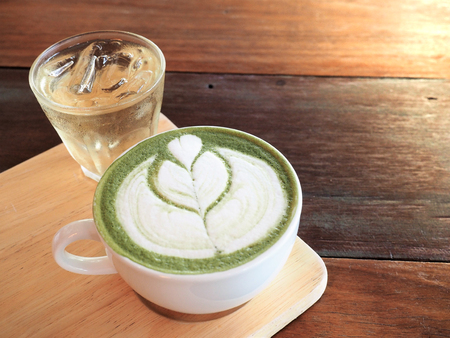 a cup of hot matcha green tea latte with latte art and a glass of water with iced serve on wooden tray.の写真素材