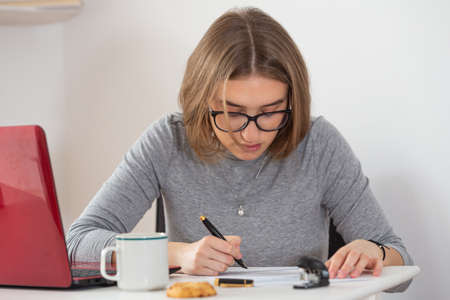 European woman writing at her desk next to the red computer. Remote work from home. Isolation time. Pandemic threat. Virus outbreak implications.の写真素材