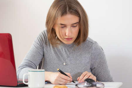 Natural blonde woman writing with her fountain pen on the sheet of paper doing remote work from home. Pandemic threat. Isolation time.Virus outbreak implications.の写真素材