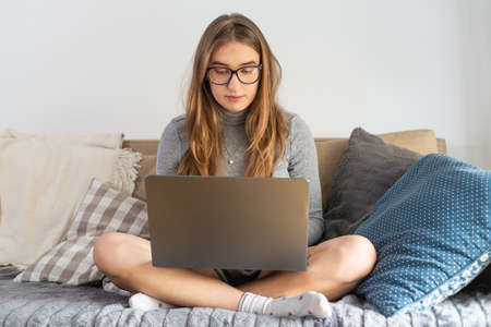 Natural blonde woman working comfortably on her laptop sitting on couch with big pillows. Remote work from home. Isolation time. Pandemic threat. Virus outbreak implications.の写真素材