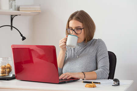 Natural blonde woman wearing gray turtleneck drinking coffee while doing remote work from home. Pandemic threat. Isolation time. Virus outbreak implications.の写真素材