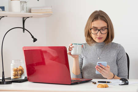 Natural blonde business woman checking her phone while drinking coffee at her desk. Remote work from home. Pandemic threat. Isolation time. Virus outbreak implications.の写真素材