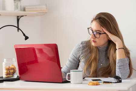 Young blonde european woman doing remote work on her red laptop in home. Isolation time. Pandemic threat. Virus outbreak implications.の写真素材