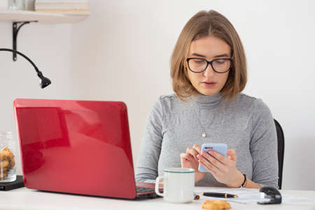 European woman with glasses checking her phone in front of the red laptop. Remote work from home. Isolation time. Pandemic threat. Virus outbreak implications.の写真素材