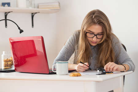 Young blonde woman with glasses writing on the sheet of paper at her white desk. Remote internet online classes. Isolation time. Pandemic threat. Virus outbreak.の写真素材