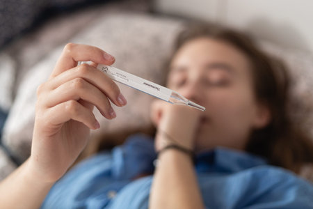 Woman holding medical thermometer while lying in bed and coughing. Coronavirus disease outbreak,