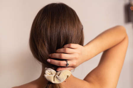 Blonde woman with beige scrunchie on her ponytail, holding it with her hand, rose ring on her finger. View from the back.の写真素材