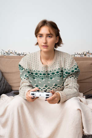 Young short haired blond woman wearing warm sweatshirt playing new video game at launch in front of tv on a couch under blanket. Front perspective, winter theme, self isolating, time at homeの写真素材