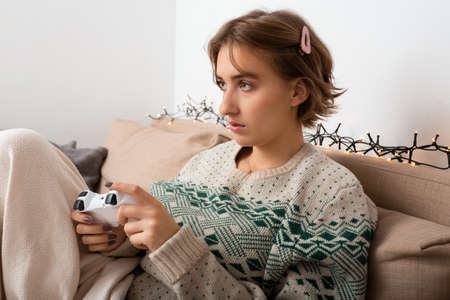 Caucasian blond woman with hair clip playing video game with controller on beige sofa wearing beige cable knit sweatshirt, cozy vibes, christmas theme lights, winter mood, isolation time, relax homeの写真素材