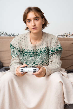Caucasian woman playing new video game on her gaming console on tv with controller sitting on couch with blanket. Cozy vibes, winter theme, warm tones, green patterned sweater, safe environment.の写真素材