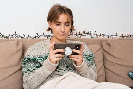 Caucasian blond girl enjoying playing game on her smart phone sitting on Ecru White couch with beige blanket. Safe and cozy space, home, warm winter cable knit sweater, christmas time, bright sceneの写真素材