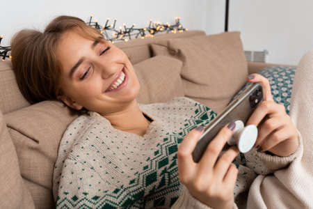 Blond young caucasian woman with perfect smile doing remote call, video conference, playing video games on her smart phone on beige couch. Warm, winter sweater, green pattern, cozy, christmas theme,の写真素材