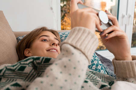 Happy blond young woman with cable knit sweater playing video games, video conference smart phone, while lying down on a comfortable sofa in home, self isolation, winter, christmas theme, warm tones,の写真素材