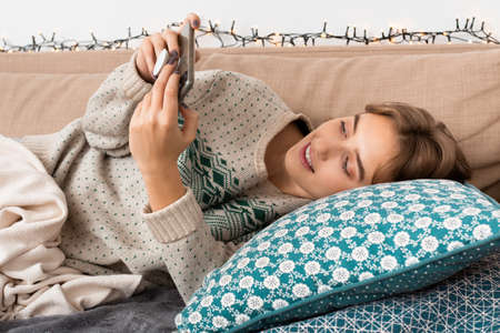 Beautiful woman with perfect smile, lying down on a sofa with christmas themed sweater watching video on her smart phone, warm tones, christmas vibes, cozy environment, short hair, beige blanketの写真素材