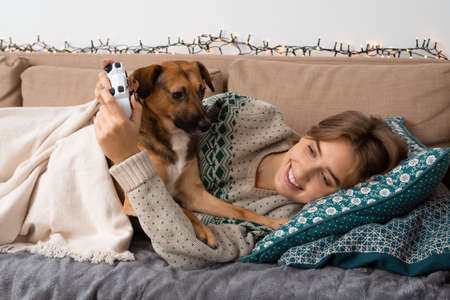 Blond caucasian woman having fun playing video games, smiling, lying on a beige sofa under blanket with her cute, mongrel dog, new launch premiere, cozy vibes, happiness, christmas time, warm sweaterの写真素材